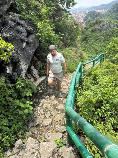 In Tam Thanh Ward, Vietnam, a man wearing a grey flat cap, a light grey t-shirt with the word "RUN" printed on it, and beige shorts walks up a stone path lined with lush green vegetation. A bright green metal railing runs alongside the path, guiding him upwards. The path is uneven, made of irregularly shaped stones, and bordered by large grey rocks and dense green foliage. In the background, a city with red-tiled roofs can be seen nestled amongst rolling green hills under a cloudy sky. The man appears to be hiking or exploring the natural landscape.