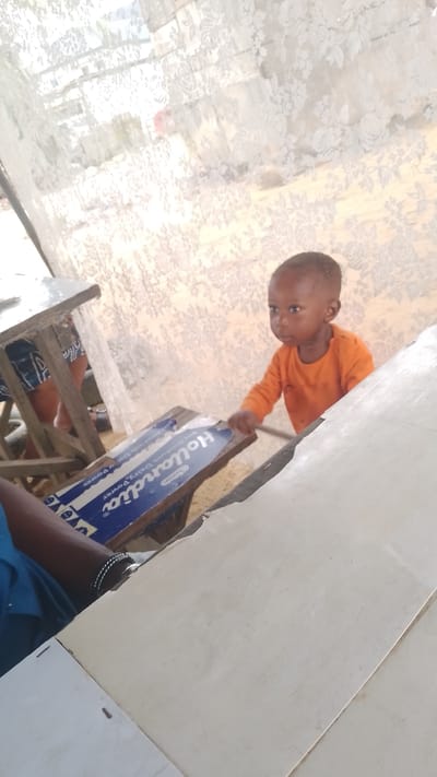 A child in an orange shirt stands next to a table in what appears to be a building in Effurun, Nigeria. The table is adjacent to one with a "Hollandia Dairy Power" advertisement attached. A person wearing a blue shirt is seated nearby with their arm and watch visible. Light filters through a patterned curtain or window covering in the background.