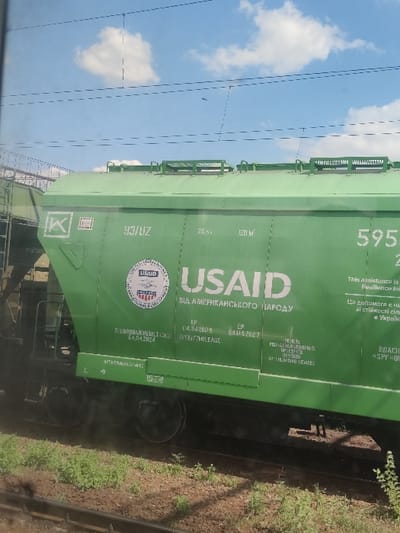 The image depicts a lime-green hopper-style railcar situated on a railway track in Fastiv, Ukraine. The railcar features prominent white lettering that reads "USAID" and, below it, "ВІД АМЕРИКАНСЬКОГО НАРОДУ" (FROM THE AMERICAN PEOPLE) in Cyrillic script. A circular USAID logo is visible to the left of the main text. Additional markings on the car include "93/UZ," numbers "70 T" and "120 M³" indicating capacity, and technical dates: "КР 04.04.2019," "ДР 04.04.2027," and "ПЕРВИННИЙ ОСМОТР 04.04.2024" (Primary Inspection 04.04.2024). Further partial text reads "This assistance is provided with Resilience In..." and "Ця допомога є част... стійкості сіл..." (This assistance is part of... resilience of villages/communities...). The numbers "595" are visible on the right side. The bottom of the railcar shows its undercarriage and wheelsets. The environment consists of a gravel ballast railway bed with sparse green vegetation, primarily weeds and grass, growing along the tracks. Above the train, a bright blue sky with scattered white clouds is visible, crisscrossed by dark overhead electrical lines, typical of an electrified railway. No individuals or specific actions are depicted in the scene.