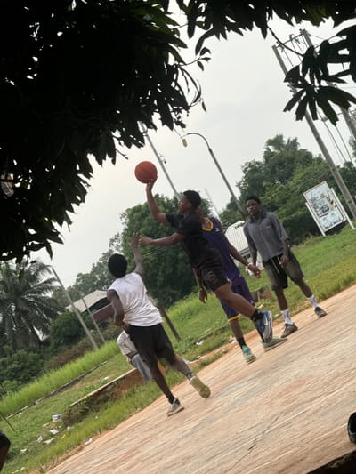 This image depicts an outdoor basketball game in progress in Amansee, Nigeria. The scene takes place on a worn, reddish-brown court with faint white lines. Four young Black men are actively playing. One individual, wearing a white t-shirt and black shorts, is jumping with his arms extended, attempting to block a shot. Another player, in a black t-shirt and black shorts with "45" visible, is in mid-air, reaching up to shoot a basketball. A third player, in a purple and yellow basketball jersey and black shorts, stands nearby, observing the play. The fourth player, dressed in a striped grey and white shirt and dark shorts, stands to the right, also watching the game.
The environment is characterized by lush green grass and vegetation bordering the court. Palm trees and other dense foliage are visible in the background, suggesting a tropical or sub-tropical setting. Overhead, the sky is overcast and appears to be a pale grey. Utility poles with electrical wires are present in the distance, along with a white building and a large white sign with text and images on it. The overall atmosphere is casual and energetic, capturing a moment of athletic activity in a semi-rural or suburban Nigerian setting.