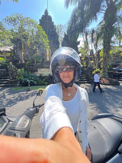 A person wearing a helmet and white clothing sits on a parked Yamaha scooter on a paved road in Kedewatan, Indonesia. The road is textured with a repeating hexagonal pattern. Beyond the road, there is lush greenery and an ornate stone structure, possibly part of a temple or historical site. A person dressed in a white shirt and dark pants is standing in the background near a parked car. The sky is blue.
