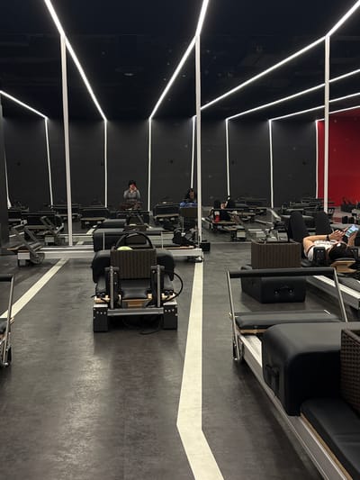 In a modern fitness studio with dark walls and ceiling, accented by linear white lighting, several pilates reformers are set up in rows on a dark grey floor marked with white lines. In the foreground, one reformer is equipped with a dark woven basket containing a yellow object. Further back, a person is lying on a reformer, looking at a mobile phone. Another person is seen in the mid-ground, taking a selfie with a phone. The studio appears to be empty of other patrons, with only a few individuals visible. The environment suggests a class or workout session is either about to begin, is ongoing, or has just concluded. There are reflections on the wall, indicating the presence of mirrors.
