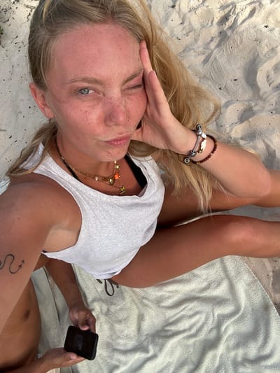 The image depicts a woman lying on a white towel on a sandy beach. She is wearing a white tank top and has blond hair. She is looking at the camera with one hand near her forehead. She is wearing bracelets on one wrist and a necklace. A man is partially visible on the right side of the frame, holding a black smartphone. He has a tattoo on his arm and is shirtless. The beach appears to be in Sainte-Anne, France.