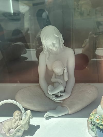 The image shows a display of porcelain figurines through a window, likely in a shop. The central figurine is a large, white porcelain sculpture of a mother cradling a baby. The mother is depicted sitting with her legs crossed, and her head is bowed, looking down at the infant. The baby is curled up in her arms, with one hand near its face. The mother's hair is long and wavy.

To the left of the central figurine, there is a smaller porcelain figurine of a baby lying in a basket with the inscription "2024". This baby is asleep, with its eyes closed and head resting on the basket's edge.

To the right of the central figurine, partially visible, is another porcelain figurine of a child holding a bouquet of flowers. The child is wearing a blue garment, and the flowers are a mix of yellow and white with green leaves.

The figurines are displayed on a white surface, possibly a shelf. Reflections are visible in the window, suggesting an indoor display with a street or building exterior outside. The lighting is bright, casting shadows from the figurines onto the white surface.

Based on the style and the presence of such decorative items, this display is likely located in Karlovy Vary, Czechia, a city known for its spa town atmosphere and its abundance of shops selling artisan crafts and souvenirs. The specific location is not precisely identifiable from the image alone, but the context suggests a retail establishment in the city center, possibly on one of the colonnades or a main street frequented by tourists.