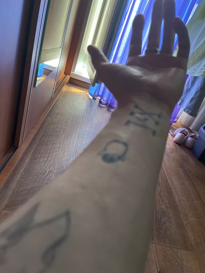 The image shows a close-up view of a person's forearm with tattoos. The tattoos appear to be Roman numerals and a circular symbol. The forearm is resting on a wooden floor. In the background, there are curtains and a pair of pink and white shoes. The lighting suggests an indoor environment, possibly a bedroom or living area. The image is taken from a first-person perspective, looking down at the arm. The location is stated to be in Muravlenko, Russia, but this information cannot be independently verified from the image itself.