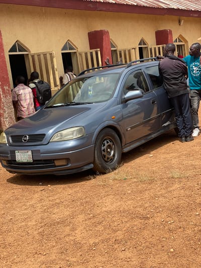 In Ban, Nigeria, a group of individuals are gathered around a dark blue Opel Astra station wagon parked on a dirt road. The car is positioned in front of a building with a yellow facade and red trim. The building has several windows with metal shutters, some of which are open, revealing dark interiors.

The individuals are primarily men. One man, wearing a light-colored plaid shirt, stands near the building, facing away from the camera. Another person, in a red shirt, is partially visible behind him. A man with a black backpack is also present. Two men are standing by the open passenger side door of the car. One is wearing a dark jacket and dark pants, and the other is in a teal t-shirt and dark pants. Their posture suggests they are engaged in a conversation or interacting with someone inside the vehicle.

The ground around the car is covered in reddish-brown dirt and small stones, with sparse dry grass growing in patches. The sky is bright, indicating it is daytime. The overall scene depicts a casual gathering or interaction related to the vehicle and the building.