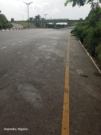 The image shows an empty asphalt road with a yellow dividing line. The road leads to a bridge in the distance. The bridge has concrete supports and is flanked by vegetation. The scene is located in Aramoko, Nigeria, at the coordinates 7.66 latitude and 5.167 longitude. There are no visible people or vehicles in the image. The surrounding environment is characterized by overgrown vegetation on either side of the road. The sky is overcast.