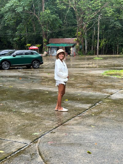 A woman is standing in a wet parking lot in front of a small building with a red umbrella and a green roof. She is wearing a white jacket, denim shorts, a hat, and white slip-on shoes. A green car is parked to her left. The background features a dense forest with tall trees. The ground is wet and shows reflections of the surroundings, indicating recent rainfall. The overall environment appears to be a natural or park-like setting, possibly near a tourist attraction or rest stop.