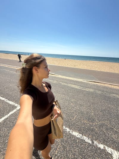 A woman is captured in a selfie on a sunny day, standing on an asphalt pathway adjacent to a sandy beach. The woman, facing away from the camera and to her right, has long brown hair styled with a green hair clip and is wearing a dark brown athletic crop top and matching shorts. She is holding a brown paper bag in her right hand. In the background, the beach stretches out towards a calm blue sea under a clear blue sky. Further in the distance, a person is walking along the beach. The pathway she is on has white painted lines. This scene is consistent with the promenade areas found along the coastline of Bournemouth, United Kingdom, likely near the town center or West Cliff area given the beach and promenade layout.