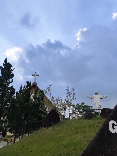 The image displays an elevated outdoor scene in Dalat, Vietnam, under a partially cloudy sky. The foreground and midground consist of a sloping, grassy hillside dotted with several large, dark, irregular rocks; one rock in the far right foreground bears a partial white letter "G". To the left midground, partially obscured by tall, dark green coniferous trees, is the Domaine de Marie Church, identifiable by its steeply pitched brown roof and a white Latin cross topped with a lightning rod. Further to the right and higher on the hill, behind a dark metal fence, stands a large, white statue of Jesus Christ with outstretched arms. The sky above is blue with scattered grey and white clouds. No people are visible.