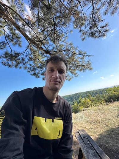 A medium shot selfie shows a man sitting outdoors with a clear blue sky and a tree in the background. He is wearing a black sweatshirt with a yellow smiley face design on it. To the right of the man, a wooden bench is partially visible. The background features a lush green forest and a valley. The image appears to have been taken in Velykozalissia, Ukraine.