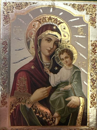 The image is a close-up of an icon depicting the Virgin Mary holding the Christ Child. The Virgin Mary is on the left, wearing a dark red robe and a burgundy veil. She is looking towards the viewer with her head tilted slightly. The Christ Child is on the right, held in her arms. He is wearing a light-colored tunic and a green cloak, and is looking to his left. Both figures have halos. The background is a pale, textured silver, with decorative gold elements in the corners and around the halos. The icon is framed by an ornate gold border with red and gold decorative patterns. There are Greek inscriptions in gold on the background next to the figures' heads. The icon itself appears to be in good condition, with no obvious signs of damage. There is no discernible information about its location in Marten, Bulgaria within the image itself.