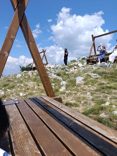 The image depicts a scenic outdoor location in Kuk, Montenegro. In the foreground, a wooden picnic table with a menu card is visible. Beyond the table, a grassy, rocky hillside stretches towards a bright blue sky with scattered white clouds. Several wooden structures, likely part of a swing set or similar recreational equipment, are positioned on the hillside.
In the middle ground, three individuals are present. A man stands on the left, wearing a black sleeveless shirt and dark pants, with sunglasses and a beard. To his right, a woman in a blue dress is seated on a wooden bench, holding a white cap to her head as if taking a photograph or shielding her eyes. Further to the right, another person in white is partially visible, also seated. The overall scene suggests a casual, recreational setting on a clear, sunny day, possibly a viewpoint or park area in the mountainous terrain of Kuk.