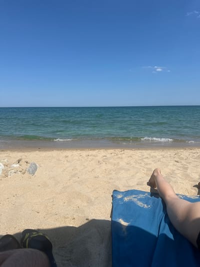 The image depicts a serene beach scene in Byala, Bulgaria, under a clear blue sky with a few scattered clouds. The foreground is dominated by golden sand, with a pair of black and yellow sandals visible on the left. In the right foreground, a person's legs are resting on a blue beach mat, with their feet bare and lightly dusted with sand. The middle ground features the gentle turquoise waves of the Black Sea lapping onto the shore, creating a shallow waterline. The horizon is a sharp line where the sea meets the sky. The overall atmosphere is one of tranquility and relaxation.