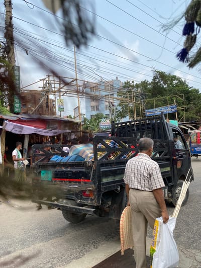 The image captures a bustling street scene in Narayanganj, Bangladesh, under an overcast sky. The primary subject is a man, seen from behind, wearing a plaid shirt and light trousers, walking alongside a black pickup truck. He carries a large white bag filled with long, thin sticks (possibly bamboo or reed) and a round, woven flat basket. The white bag has Bengali text "খাবার পরিবেশনায়" (Khabar Poribeshonay), meaning "In food serving/presentation."

The black truck, possibly a Mahindra based on a faint visible logo, has a cage-like rear and appears to be stopped or moving slowly on the road. Its green Bangladeshi license plate "ঢাকা মেট্রো ন-১৫-০০৮২" (Dhaka Metro N-15-0082) is visible at the rear. Inside the truck's cabin, another person can be seen.

The street is characterized by numerous overhead electrical wires and an elaborate network of bamboo scaffolding, which likely supports temporary market stalls, advertisements, or construction. Several banners, one featuring a political figure or local leader and Bengali text, are hung from these structures. A blue sign visible on the scaffolding reads "রাবিব স্টোর" (Rabib Store) and "রাবিব ট্রেডার্স" (Rabib Traders), along with contact numbers. Another green sign below the power lines says "নিউ পপুলার ফার্নিচার" (New Popular Furniture).

In the mid-ground, near the scaffolding, another person is visible, possibly a street vendor. The overall impression is one of everyday activity in an urban or semi-urban South Asian setting, with elements of commerce and local infrastructure. The time of day appears to be daytime, given the light, though the overcast sky creates a diffused light.