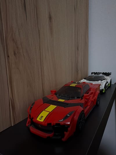 Two toy race cars are displayed on a black shelf. The car in the foreground is red with yellow racing stripes and a small yellow emblem on the hood. The second car, behind and to the right of the first, is white with green accents. The cars are positioned in front of a light brown wooden textured surface that appears to be a wall or furniture. The overall lighting suggests an indoor setting. There is no discernible information in the image that would indicate a specific location in Sankt Augustin, Germany.