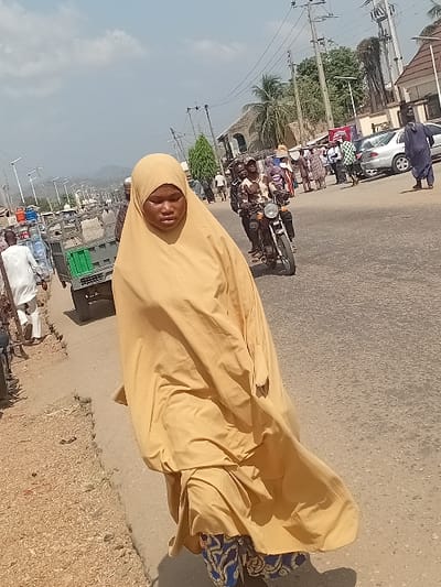 A woman in a tan hijab stands on a road. A motorcycle carrying multiple passengers moves past. Cars, trucks, and people line the street beyond. A building with a rounded roof and nearby palm tree is also visible. Utility poles run along the side of the road. The sky is pale blue. The image is rotated sideways.