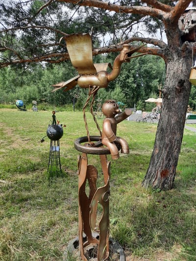 In Зеленогорский, Russia, an outdoor sculpture garden features several whimsical metal artworks. In the foreground, a large metal sculpture depicts a bird with outstretched wings perched above a circular frame supported by abstract human-like figures. To the left, another sculpture shows a small figure resembling "The Little Prince" standing on a black sphere, which is itself mounted on a wire stand. In the background, a blue globe sculpture is visible, along with other indistinct metal art pieces and trees. The sculptures are situated in a grassy area with a large pine tree dominating the right side of the frame.