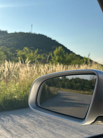 The image captures a view from a moving vehicle, focusing on a side-view mirror reflecting a winding road through a wooded area. In the background, a grassy hillside is visible under a clear blue sky. A tall communication tower stands on the crest of the hill. The foreground shows the car's exterior and the road surface, with long shadows suggesting it is late afternoon or early morning. The scene appears to be located on the outskirts of Prisovo, Bulgaria, given the landscape and vegetation.
