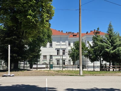 The image depicts an outdoor scene, likely a sports court, in front of a pale blue building with a red tiled roof. The building appears to be a school or a similar institution, characterized by its numerous windows and architectural details. A tall, grey utility pole stands prominently in the foreground, to the right of a large tree with dark green foliage. A white pole, possibly part of a sports net, is visible on the left side of the image, with a white object lying at its base. A chain-link fence separates the sports court from the building and the surrounding grounds, which include more trees and some picnic tables to the right. The sky is clear and blue. The scene is captured on a sunny day, casting distinct shadows on the court. The location is identified as Lom, Bulgaria.