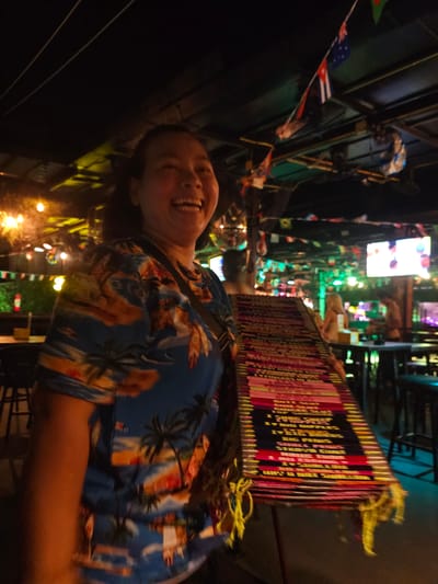 This is a slightly blurry, low-angle, close-up, outdoor, nighttime photograph of a person. The person, who appears to be a woman with a broad smile and teeth showing, is wearing a blue Hawaiian shirt with a pattern of palm trees and a beach scene. She is looking directly at the camera. The scene behind her is a busy, lit-up street or market, likely in Thailand, given the context. Rows of stalls or shops are visible, with colorful displays and bright lights. Some flags, possibly national flags, are hanging from poles in the background. A prominent object in the mid-ground is a stack of what appears to be books or framed items, with spines showing text. The lighting is a mix of artificial, colorful lights from the stalls and streetlights, indicating it is nighttime. The overall atmosphere is lively and festive. The subject's expression conveys happiness and excitement. The image is slightly tilted, adding to the dynamic feel.
