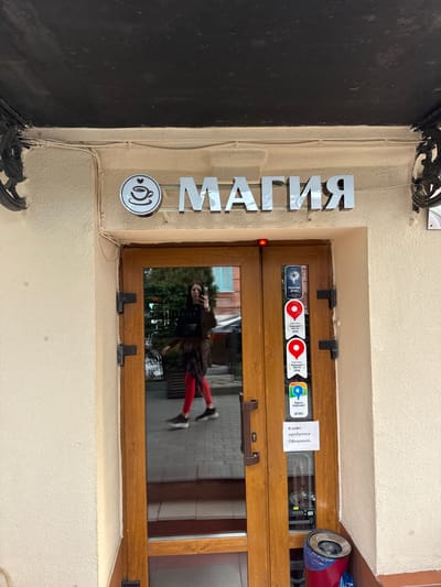 Here's a detailed description of the image:
**Scene:** The image is a view of a shop entrance, possibly a cafe called "МАГИЯ" (Magia or Magic in Russian).
**Location:** While the OCR text mentions that the cafe is a "Good place 2GIS", Akhieli is not mentioned in the OCR.
**Objects:**
* A brown wooden double door is visible with a small window.
* Stickers with the logos "2GIS" and "Yandex Maps" are on the window.
* There is a sign indicating that the cafe needs a waiter.
* The shop name "МАГИЯ" is displayed vertically on the side of the doorframe in silver lettering.
* There is a small round sign with an image of a coffee cup next to the name.
* A security camera is visible above the door.
* A black wrought iron design is visible on the left side.
**Reflection:**
* A person's reflection can be seen in the window, wearing a dark coat, and bright red leggings. They are holding a phone, possibly taking a picture.
**Environment:**
* The building exterior is a light beige color.
* The sidewalk is visible through the reflection in the window.
**Overall Impression:** The image is likely taken in a public area, showing a cafe entrance with various informational signs and a person reflected in the window.