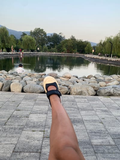 The image shows a paved area in Lagodekhi, Georgia, with a pond in the background. A person's leg, wearing a sandal, is in the foreground, stretched out towards the pond. Several people are visible near the edge of the pond, with some appearing to interact with the water. The pond is surrounded by large rocks, and beyond that, there are trees and hills. The sky is clear and light.