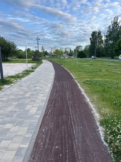 The image depicts a paved pathway and a reddish-brown surface, likely a running or cycling track, in the city of Kommunar, Russia. The pathway on the left is made of gray interlocking pavers arranged in a patterned fashion. Adjacent to it on the right is the darker track. Beyond the track, a grassy area with small white flowers stretches towards a road. Trees line the background, with some buildings visible in the distance, including a taller white building. The sky is partly cloudy with a mix of blue sky and gray clouds. There are no people or specific actions clearly visible in the scene, making it a neutral depiction of an outdoor public space.