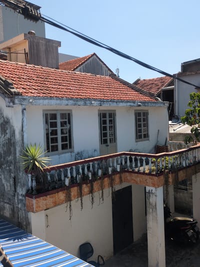 The image shows a building exterior in Tuy Hòa, Vietnam. The building has a red tile roof, a white facade, and a concrete railing with decorative white balusters and a red-brown tiled top. There are three windows on the facade, each with multiple panes. A spiky plant grows near the railing on the lower right. To the right of the building is an open area with a dark door, a white pillar, and a parked scooter in the background. A blue and white striped awning is visible at the bottom right. Power lines are seen against the blue sky in the upper left.