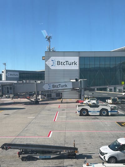 The image shows an airport tarmac in Arnavutköy, Turkey, on a clear, sunny day. A jet bridge, labeled with "BtcTurk," connects to a terminal building also bearing the "BtcTurk" logo. Two airport tow tractors are visible on the tarmac. One is a white tractor with blue accents and the "Çelebi" logo, positioned in the foreground. Another similar tractor is further back. A baggage conveyor belt is seen on the left side of the frame, with a fire extinguisher attached. A white passenger vehicle with "A-03" markings is parked on the right. The tarmac is marked with red and white lines. In the background, the modern terminal building features large glass windows, and a communication tower with antennas is present. A single individual wearing an orange vest is walking on the tarmac near the jet bridge.