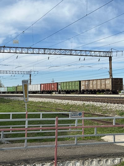The image shows a railway scene in Кечёво, Russia. A train consisting of multiple freight cars of varying colors (brown, green, and white) is on the tracks. Overhead, there are power lines supported by a metal structure. A pedestrian crossing with a metal barrier and signage is in the foreground. One sign reads "POST" vertically. The other is a directional sign for crossing. The sky is visible in the background with scattered clouds.
