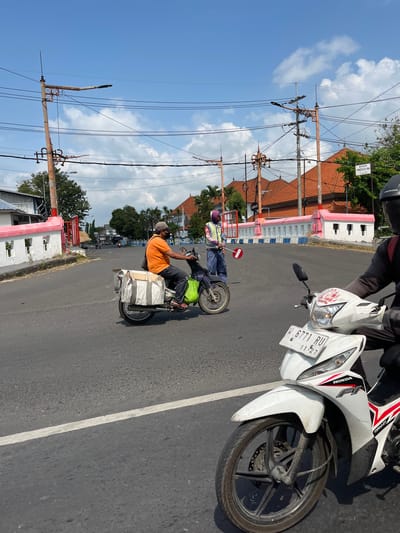 In Kludan, Indonesia, on a sunny day with a clear blue sky and scattered clouds, a street scene unfolds. The road, marked with a white line, features several motorcycles. In the foreground, a white Honda motorcycle, with license plate W 6771 RU, is prominently displayed, its front wheel and part of the body visible. To the left, another motorcycle is seen carrying a rider in an orange shirt, dark pants, and a cap, with a large bag strapped to the back. Further down the road, a traffic officer in a vest and helmet is directing traffic with a stop sign. Behind the officer, a building with a pink and white facade and terracotta tiled roofs stands. Overhead, electrical poles and wires crisscross the sky, common infrastructure in urban areas. A sign that reads "BIDAN YUNIS R. S. Tg. Keb" is visible on a pole. The overall environment suggests a typical day in a bustling Indonesian town.