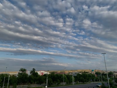 The image captures a wide-angle view of a cityscape under a dramatic sky filled with layered, textured clouds. The sky dominates the upper half of the frame, with streaks of blue visible between the gray and white cloud formations.
In the foreground, a road runs horizontally across the lower portion of the image, with a few vehicles visible, including a white car. Streetlights and traffic signs are present along the roadside. A line of trees runs along the edge of the road, creating a green buffer.
Beyond the trees, various buildings are visible. To the left, a modern building with red accents is prominent. Further to the right, a cluster of buildings with red roofs is nestled amongst more trees. In the mid-ground, a distinctive Islamic-style building with a turquoise dome and two minarets stands out against the landscape, likely a mosque. This structure appears to be located within the city of Zaamin, Uzbekistan, given its architectural style and typical setting. The background consists of rolling hills and distant mountains, suggesting a semi-arid or mountainous region. The overall scene is an urban landscape integrated with natural elements, under a dynamic sky.