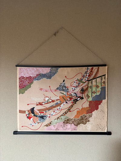 A rectangular artwork, resembling a traditional Japanese ukiyo-e style print or textile, is displayed on a light brown, finely textured wall in Nozawaonsen, Japan. The artwork depicts multiple stylized figures in elaborate traditional Japanese attire, arranged diagonally across the frame. These figures are interwoven with numerous flowing, patterned ribbons in various colors such as red, blue, green, orange, pink, and white. Red lines connect elements throughout the composition. The background of the artwork is a neutral light brown, against which several large, cloud-like or abstract landscape forms in complementary patterns and colors are depicted. The top and bottom edges of the artwork are clamped by thin, black horizontal bars. A light-colored string forms an isosceles triangle, connecting the upper bar to a small, metallic hook anchored in the wall at the apex of the triangle. The wall exhibits a consistent, granular texture.