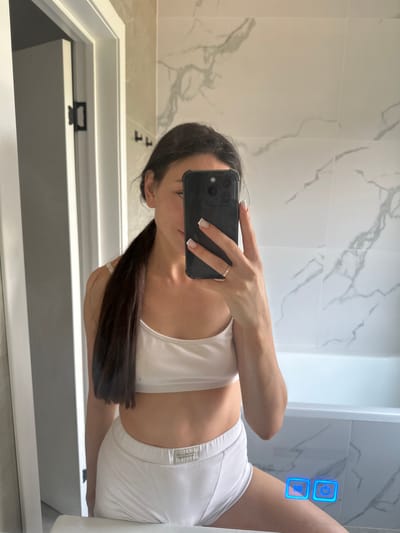The image is a mirror selfie taken in a bathroom. A young woman with dark hair pulled into a ponytail is wearing a white sports bra and white underwear. She is holding a smartphone in her right hand, which is obscuring part of her face. The bathroom features marble-patterned tiles on the wall and a white bathtub. There are also two illuminated blue buttons on the mirror, one showing "100" and the other a power icon. The location is identified as Estachi, Russia, but this information is not visually present in the image itself.