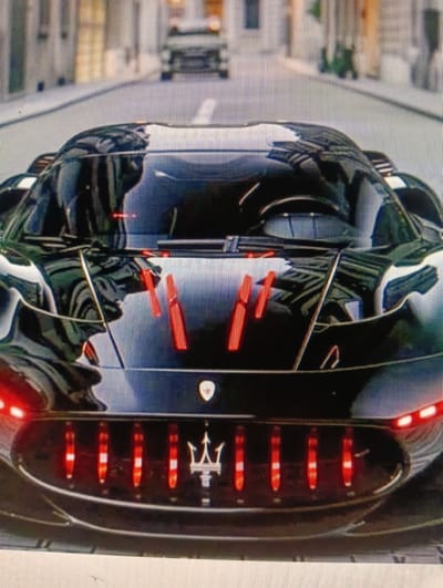 The image shows the front of a black Maserati MC20 car. The car has red vertical accents on the hood and red taillights arranged vertically within a black grille. The Maserati trident logo is visible on the grille. The car is parked on a street that appears to be in a city. The background is out of focus but shows buildings and other vehicles. There are no people visible in the image. The scene is a close-up shot of the car's front.