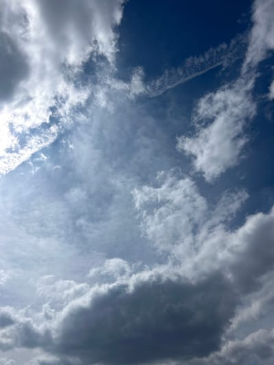 A vertical photograph captures a mixed cloudscape over Nepraznentsi, Bulgaria. The sky transitions from a deep, clear blue in the upper right quadrant to lighter blues and various shades of grey and white clouds across the remainder of the frame. Brightly illuminated, fluffy white cumulus clouds are prominent on the left side, particularly in the upper left. A distinct, linear white contrail traverses the upper right portion of the deep blue sky, extending diagonally from top right towards the center. The lower half of the image is dominated by darker, denser grey clouds, indicative of thicker cloud cover. Scattered, wispy white clouds are interspersed throughout the mid-ground. No human subjects, man-made structures, or ground features are visible within the frame.