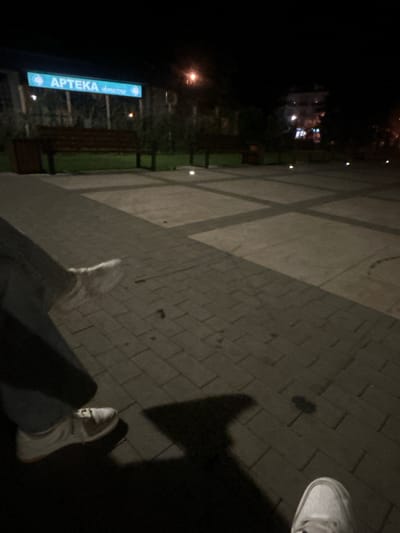 The image depicts a nighttime scene in an outdoor urban setting. In the background, a brightly lit sign reads "APTEKA Słoneczna," identifying it as a pharmacy in Siedlce, Poland. The area appears to be a public space with paved walkways and dark-colored benches. In the foreground, the lower legs and feet of at least two individuals are visible, one on the left wearing blue jeans and white sneakers, and another on the right wearing white sneakers. The lighting suggests artificial illumination from streetlights or nearby buildings, casting shadows on the ground. The overall atmosphere is dark and somewhat blurry, characteristic of a low-light photograph.