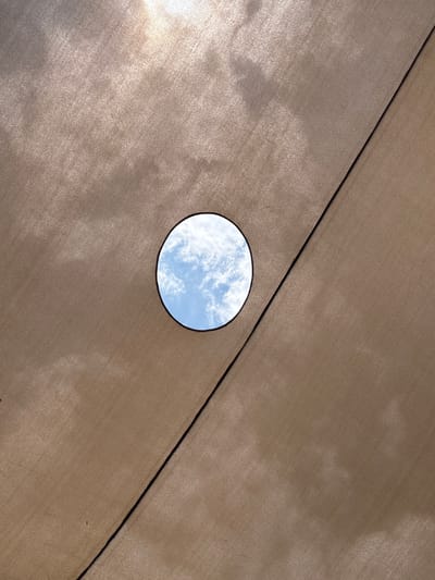 The image is a view looking up at a beige fabric sunshade, possibly part of an awning or canopy. The fabric has a textured surface. A circular hole is cut out of the fabric, revealing a patch of blue sky with white clouds. A black seam bisects the shade. Based on the location mentioned, the image could be from a public space, a residential area, or a commercial area in Or Yehuda, Israel. No people are visible.