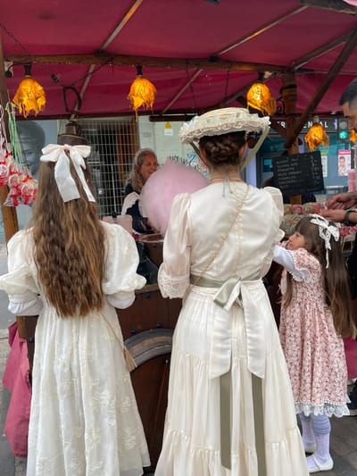 This image captures a scene at an outdoor market or fair, possibly in Terrassa, Spain. Three girls are wearing old-fashioned dresses with lace and ribbons, which are indicative of a historical reenactment or special event. One girl wears a white bonnet. An elderly woman with a gray headband holds a large pink cotton candy, while a vendor's stall is visible with lanterns hanging above and a menu board listing prices for items like "Algodon Palo" (cotton candy on a stick), "Piruletas" (lollipops), and "Manzanas" (apples), with the option to pay via "Bizum." Lollipops are also visible on display. The stall has a red awning, and parts of street signs and building facades are visible in the background.