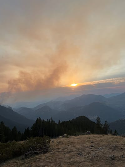 The image captures a mountain landscape, likely in or near San Juan de Plan, Spain, under a sky filled with smoke from a wildfire. The sun is partially obscured by the smoke, casting a warm, orange glow across the sky and the distant mountain ranges. The mountains themselves are layered, with the closest ones in shadow and the furthest fading into a hazy blue. In the foreground, dry, golden grass covers the ground, with scattered rocks and a few low-lying shrubs. A dense line of dark green pine trees runs along a ridge in the middle ground, with a few taller trees interspersed. The smoke in the sky appears to be originating from the left side of the frame, with plumes rising and drifting eastward. There are no people or animals visible in the image, and no specific structures are clearly identifiable, although a faint suggestion of a village can be seen in the far distance on the right. The overall scene evokes a sense of dramatic natural beauty tinged with the somber reality of a wildfire.