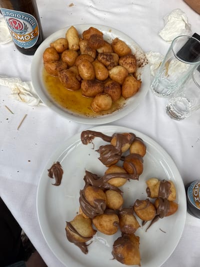 The image provides an overhead view of a dining table located in Nea Smirni, Greece. The table is covered with a white tablecloth. Two white ceramic plates of Loukoumades (Greek donuts) are centrally positioned. The upper plate contains approximately 25-30 golden-brown fried dough balls partially submerged in a golden-brown syrup, likely honey, with visible cinnamon dusting. A brown glass bottle, labeled 'ΜΠΕΛΟΠΟΙΟΤΗΣ ΣΑΜΟΣ' (Peloponnesian Samos), stands to the upper left of this plate. To the right of the upper plate are two clear glass tumblers of differing heights and several crumpled white paper napkins. The lower plate holds approximately 20-25 similar golden-brown fried dough balls extensively covered with a thick, dark brown, chocolate-hazelnut spread. A dark-colored beverage container with a partial blue label is visible to the right of the lower plate. Two small wooden toothpicks and minor food debris are scattered on the tablecloth, indicating recent consumption. No people are visible.