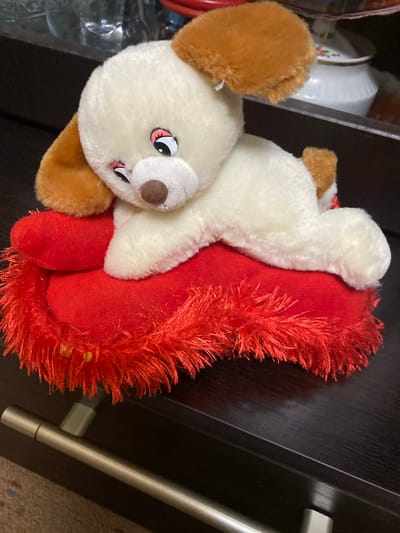 A white and brown plush dog toy lies on a red heart-shaped pillow with red fringe. The toy dog has large, dark eyes with pink accents, a brown nose, and floppy brown ears. The pillow is textured and appears to be made of velvet. The scene is set on a dark, glossy wooden surface, likely a piece of furniture. In the background, partially obscured, are shelves containing various items, including glass bottles and a white ceramic container with a floral pattern, suggesting an indoor domestic setting. A silver drawer pull is visible in the lower part of the image. The lighting is dim, casting soft shadows. This image does not provide any specific information to locate it within Yokohama, Japan.