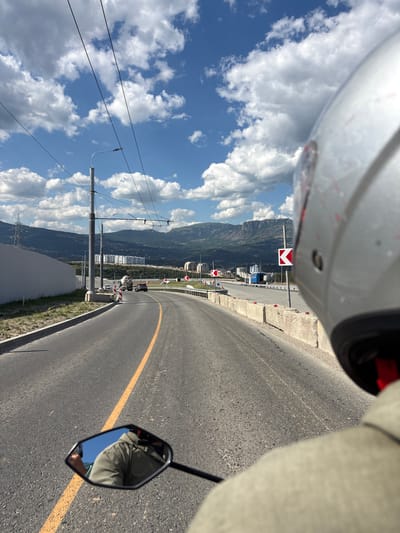 A first-person perspective captures a view from a motorcycle on a paved road under a bright blue sky with scattered white clouds. The road curves to the right, marked by a solid yellow line. In the foreground, the rider's silver helmet and part of their shoulder are visible. A motorcycle rearview mirror reflects the rider's head and shoulders, wearing a green jacket, and a bright blue sky.
The road leads towards a city nestled between mountains in the distance. Buildings are visible on the slopes and at the base of the mountains. To the left of the road, there is a low, light-colored wall. To the right, a concrete barrier runs alongside the road, with traffic signs indicating a sharp turn to the left. Several vehicles, including a cement mixer truck and cars, are on the road ahead. Overhead, power lines are strung between tall poles. The scene suggests a journey on the outskirts of a city, possibly on a highway or a major road leading into an urban area.