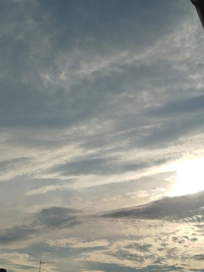 The image displays a cloudy sky over Rozniszew, Poland. The sky is a mix of gray and white clouds, with streaks of lighter clouds indicating wind patterns. The sun is partially obscured by clouds in the upper right portion of the image, casting a bright glow. In the lower part of the image, a portion of a building is visible on the left, and a television antenna stands on a pole, extending upwards. The overall scene depicts an outdoor, daytime environment under a cloudy sky.