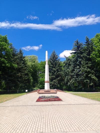 The image depicts a war memorial with a tall, obelisk-shaped monument in the center. The monument is white with some red and gold detailing, and appears to have an inscription or ornamentation at its base, though it is not clearly legible. In front of the monument is a flower bed with red and white flowers, and a wreath. The area around the monument is paved with light-colored rectangular stones, forming a symmetrical pattern. Flanking the paved area and the monument are grassy lawns and a dense line of evergreen trees, predominantly fir trees, with some deciduous trees interspersed. The sky above is a bright blue with scattered white, wispy clouds. The scene is set outdoors on a sunny day. Based on visual cues commonly associated with such memorials, and potentially correlating with other information if available, this could be a war memorial located in Yessentukskaya, Russia, likely commemorating fallen soldiers.