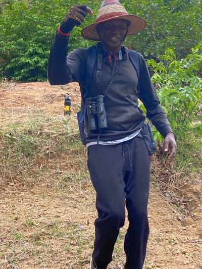 A man wearing a straw hat, black sweatshirt, black pants, and gloves is standing on a patch of dirt with dry grass and some green vegetation. Binoculars are slung around his neck, and he appears to be holding a small electronic device. The background includes green bushes and trees. The location is presumed to be Ganawuri, Nigeria.