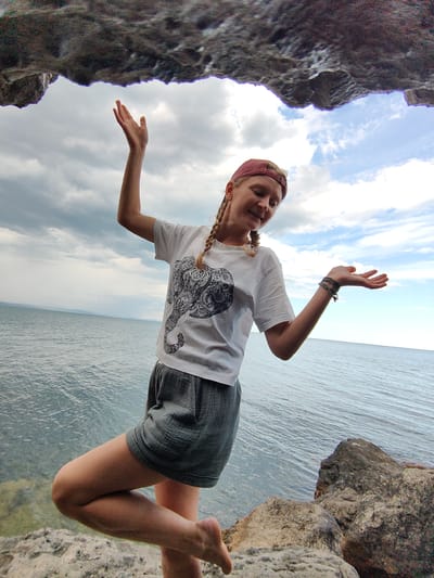The image depicts a woman standing on rocky terrain near the sea, with a dramatic rock overhang framing the top of the scene. She is wearing a white t-shirt with an elephant print, grey shorts, and a red baseball cap. Her hair is styled in two braids. She is posing with her arms outstretched, her right arm reaching upwards and her left arm extended outwards with an open palm. The sea stretches out before her under a cloudy sky. The location is identified as Utios, Ukraine.