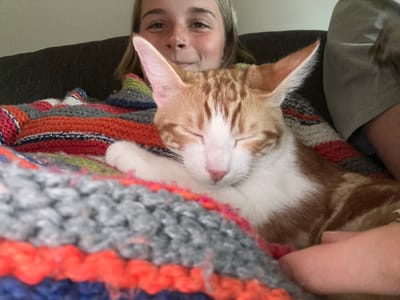 A horizontally oriented image shows a female and an orange and white cat laying under a colorful crocheted blanket. The woman's face is visible at the bottom of the image. She has fair skin, freckles, and light-colored eyes. The cat, partially obscured by the blanket, is positioned above the woman, appearing to rest on her. The cat's eyes are closed. The blanket is multi-colored with orange, gray, blue, white, and green stripes. The scene suggests a cozy, indoor setting. The city of Squamish, Canada, is not discernible from the image.