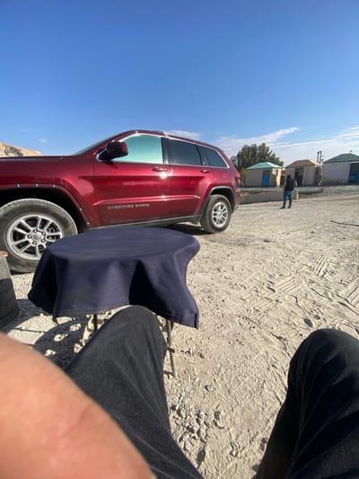 A red Jeep Grand Cherokee is parked on a dusty, possibly gravel-covered surface. A person wearing dark jeans and sitting in a chair is visible in the foreground. A dark blue seat cover or item of clothing rests on the chair. In the distance, there are a few small buildings and what appears to be a person walking. The sky is clear and blue. The image is taken from a low angle.